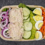 Bowl of salad with tuna salad, cucumber slices, red onion, hard-boiled eggs, shredded carrots, and greens.