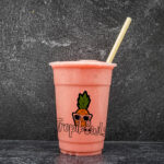 Pink smoothie cup with a straw and a pineapple wearing sunglasses on a dark textured background, tropical-themed design visible on the cup area.