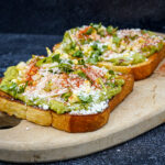 Two slices of avocado toast on a wooden board, topped with corn, herbs, cheese, and red chili flakes.
