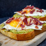 Toast topped with mashed avocado, a sunny-side-up egg, and crisp bacon pieces on a wooden board at the table edge.