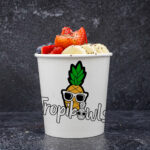 Cup of frozen yogurt topped with strawberries, bananas, blueberries and sesame seeds, with a pineapple mascot wearing sunglasses on the cup