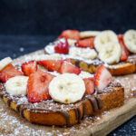 Toast slathered with chocolate spread, topped with banana and strawberry slices on a wooden board dusted with powdered sugar." It should be concise