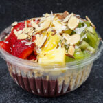 Fruit parfait in a clear cup topped with sliced almonds, coconut flakes, strawberries, pineapple and kiwi pieces on a dark countertop.