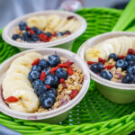 Three smoothie bowls topped with banana slices, blueberries, goji berries, and granola on a bright green woven basket/surface.