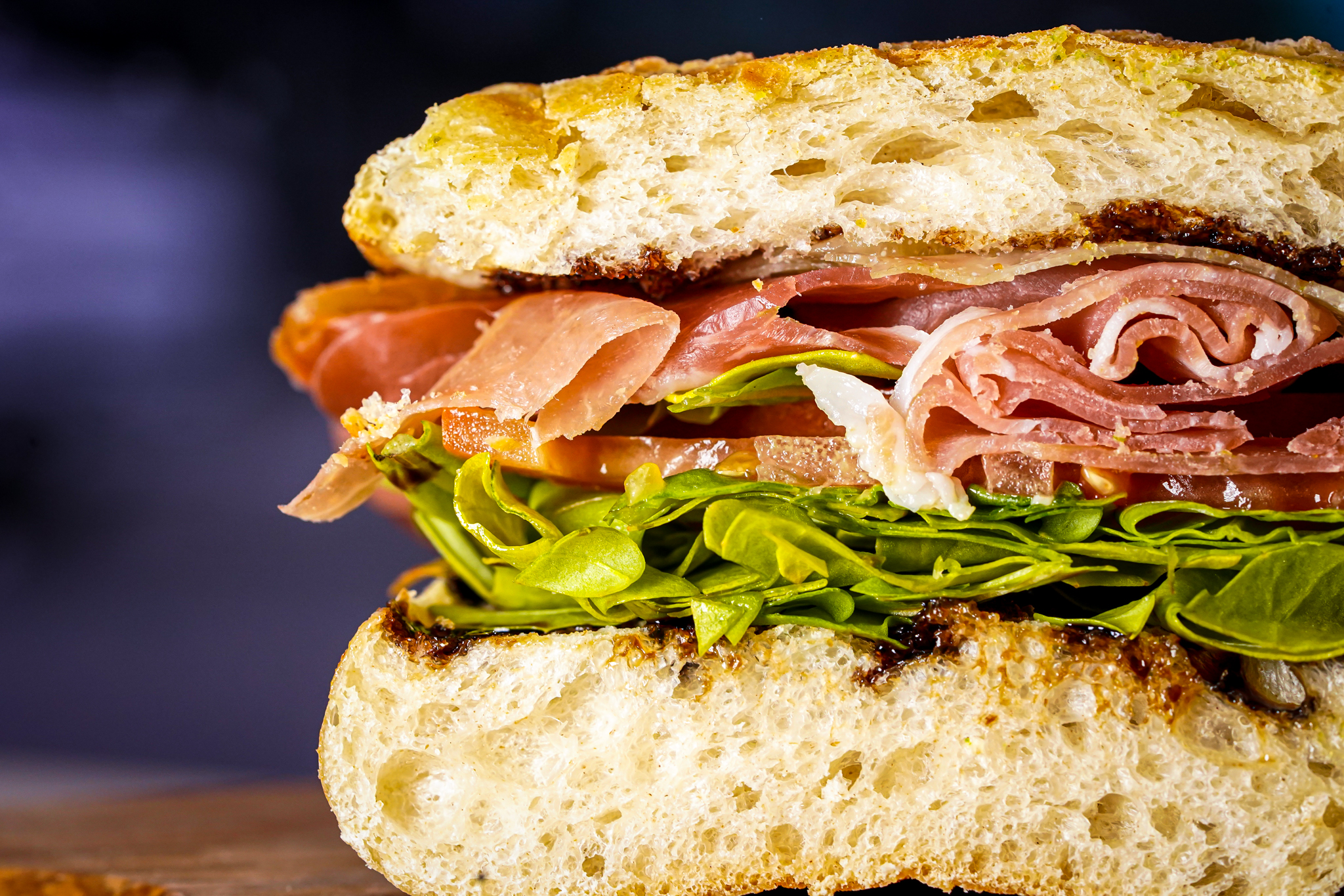 Close-up of a stacked sandwich with rustic bread, thinly sliced ham, melted cheese, and leafy greens.