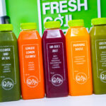 Five GOJI juice bottles in green, orange and amber tones lined up, with labels listing ingredients like cucumber, apple, celery, spinach, ginger, beet and kale.