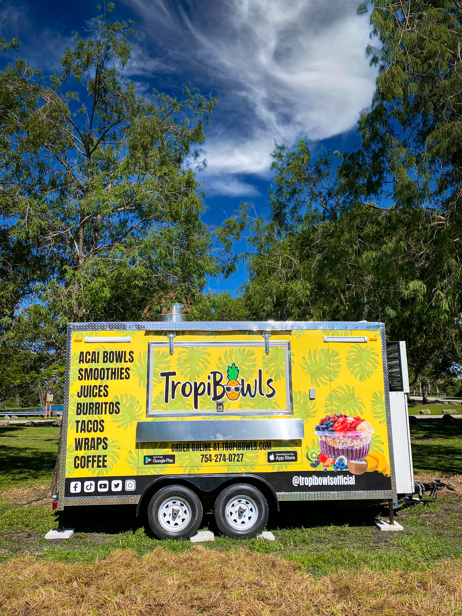 Bright yellow TropiBowls food trailer with tropical graphics, showing menu items like acai bowls, smoothies, juices, burritos, tacos on a sunny day under trees.