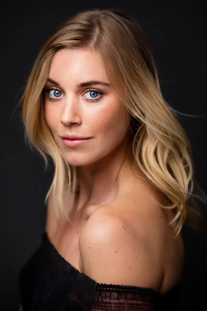 Close-up portrait of a blonde woman with blue eyes, wearing an off-shoulder black top, against a dark background.