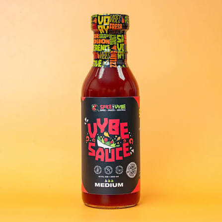 Bottle of Vybe Sauce with a black label and colorful graphics, labeled Medium, on a yellow background.