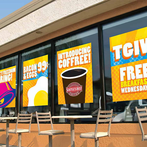 Cafe storefront with large orange promotional posters in the windows and outdoor chairs along the sidewalk.