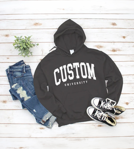 Casual outfit laid out on a white wooden floor: black hoodie with 'CUSTOM University' text, ripped blue jeans, black-and-white sneakers, and a small potted plant.