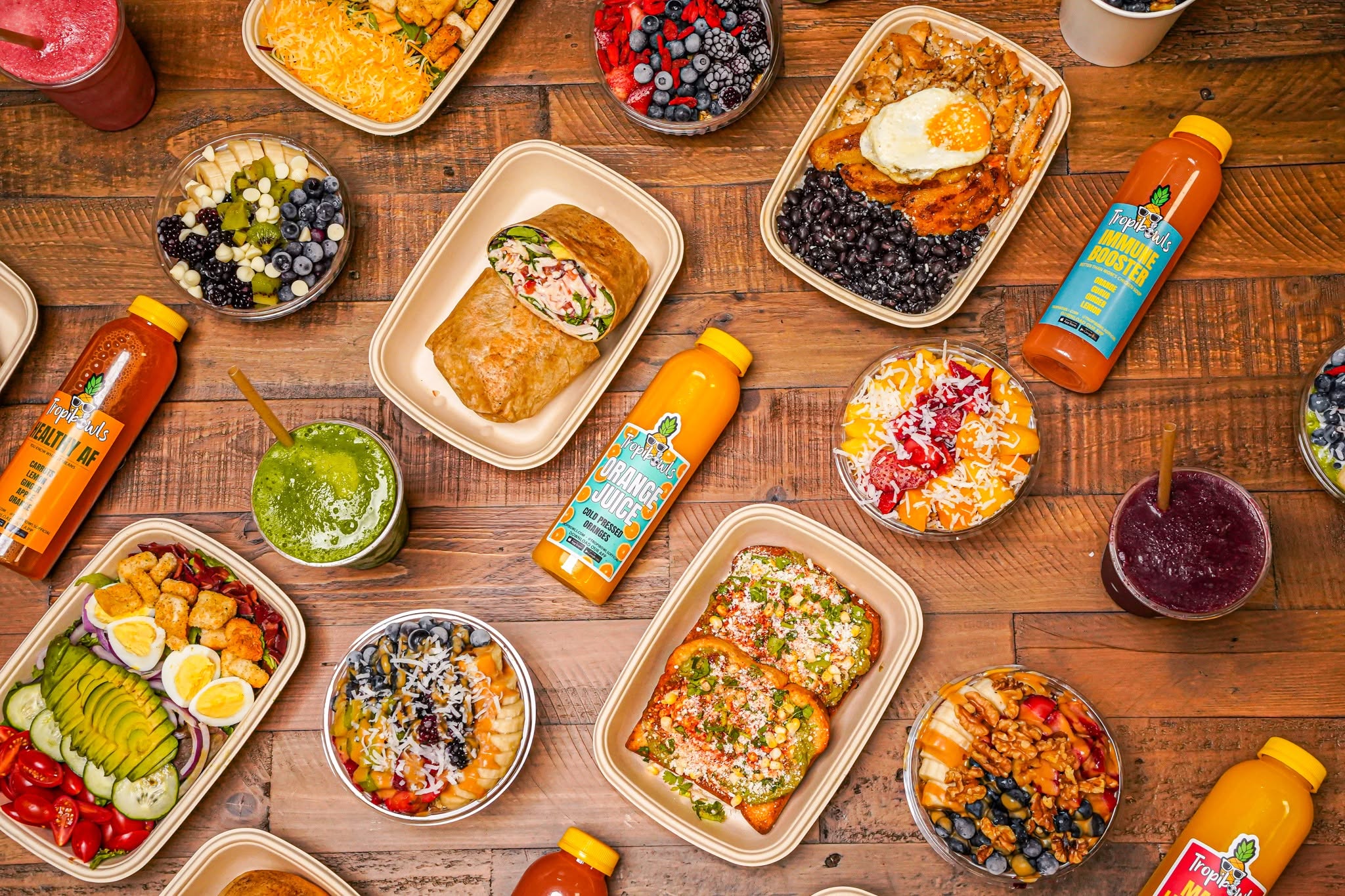 Overhead view of a wooden table with meal-prep containers of burritos, egg dishes, beans, and fruit bowls, plus smoothies and juice bottles.