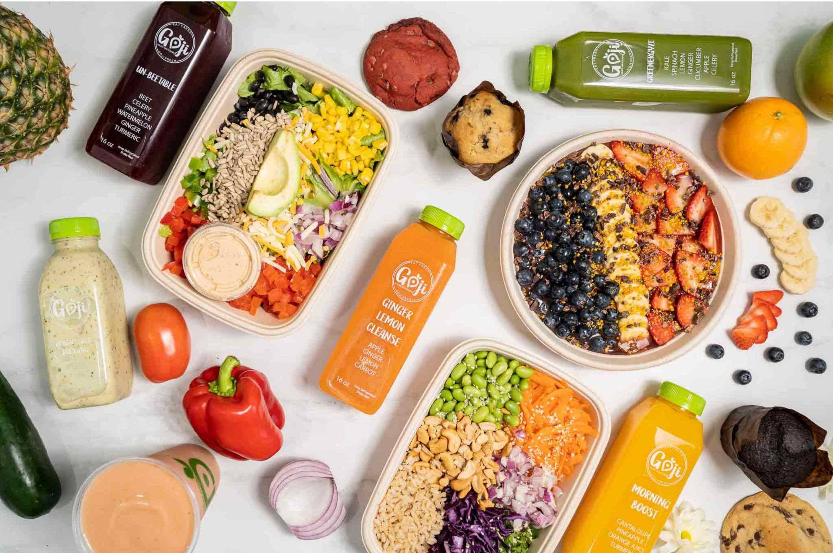 Top-down view of colorful health foods: two meal-prep bowls with veggies, avocado, and seeds, plus GOJI juice bottles and assorted fruits.