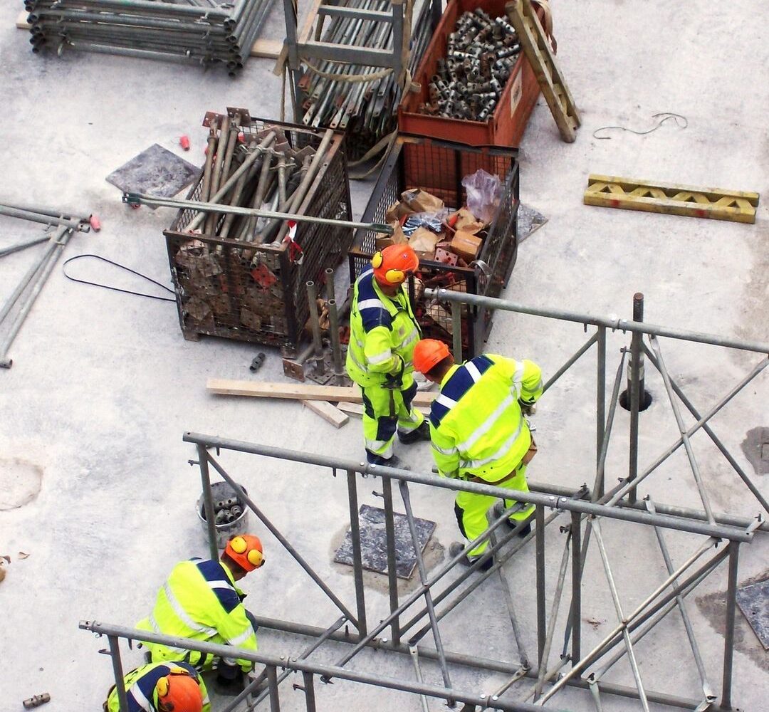 Construction workers in high-visibility outfits and orange hard hats assemble metal scaffolding on a concrete site with crates, pipes, and tools nearby.