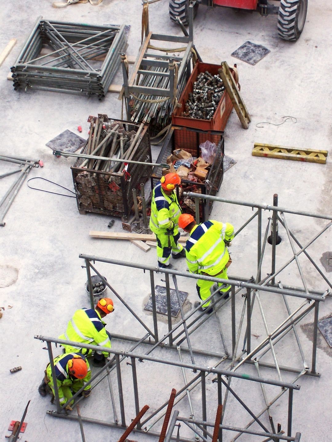 Construction workers in high-visibility outfits and orange hard hats assemble metal scaffolding on a concrete site with crates, pipes, and tools nearby.