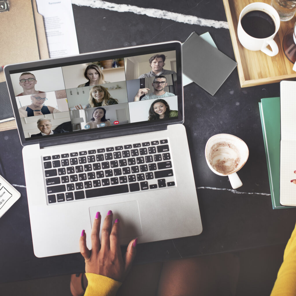 Laptop screen shows a grid-style video conference with multiple participants; foreground hands in a yellow sweater rest on the laptop keyboard, with coffee cups and desk items nearby.