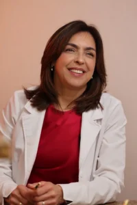 Smiling female doctor in a white coat and red blouse, seated indoors.