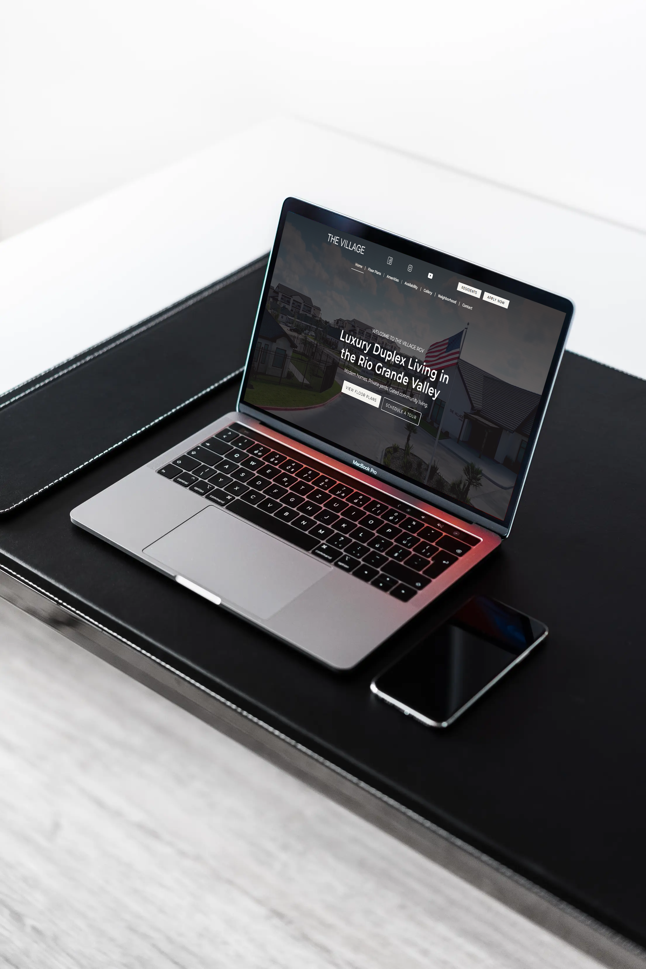 Laptop on a black desk displaying a real estate website for 'The Village' with a hero image of homes and the headline 'Luxury Duplex Living in the Rio Grande Valley' next to a smartphone on the desk surface