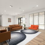 Modern lobby seating with brown leather sofas and orange chairs on a geometric rug, large glass doors, and natural light.