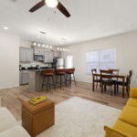 Open-concept living area with a kitchen island, gray cabinets, and pendant lights above the counter; dining table nearby and a bright yellow sofa on a light rug.