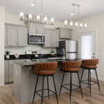 Modern kitchen with gray cabinets, stainless steel appliances, and a central island with brown tufted bar stools under glass pendant lights from above all around a breakfast bar.