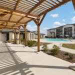 Wooden pergola over a concrete patio beside a sparkling pool at a sunny apartment complex.