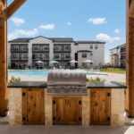 Outdoor stainless steel barbecue grill built into a stone and wood cabinet under a wooden pergola, with a pool and modern apartment buildings in the background.