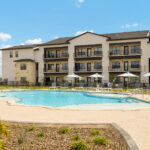 Apartment complex with balconies surrounding a clear blue swimming pool on a sunny day.