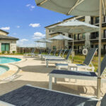 Poolside area with lounge chairs and large umbrellas beside a courtyard hotel building under a blue sky