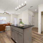 Open-concept kitchen and living room with a gray island, pendant lights, and brown leather seating in soft beige walls and wood-look tile floors.