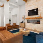 Modern living room featuring a stone fireplace with a mounted TV above a wooden mantel, beige walls, and brown leather sofas facing a round wooden coffee table.