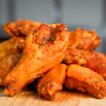 Pile of spicy fried chicken wings stacked on a wooden board, glossy with seasoned, crispy coating.