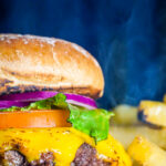 Close-up of a cheeseburger with melted cheddar, lettuce, tomato and red onion on a toasted bun, with fries in the background under a blue backdrop.