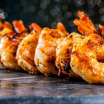 Close-up of grilled shrimp skewers lined up on a wooden surface, glossy orange glaze and char marks with a blurred bokeh background.