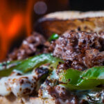 Close-up of chopped grilled beef and green bell peppers on a toasted bun, with a warm fire in the background.
