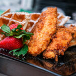 Crispy fried chicken with powdered-sugar waffles and a fresh strawberry on a dark plate.