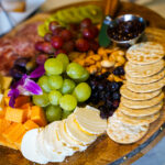 Round wooden charcuterie board with cheese cubes, green and red grapes, crackers, nuts, dried fruit, and a metal bowl of jam.