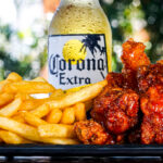 Pile of golden french fries and sticky BBQ-style chicken bites with a Corona Extra bottle blurred in the background.