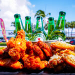 Pile of crispy fried chicken in the foreground with green Heineken bottles in a sunny outdoor setting behind.