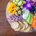 Round charcuterie board with cheese cubes, olives, grapes, salami, crackers, carrot and celery sticks, nuts, and a purple orchid centerpiece on a wooden table.