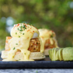 Crostini topped with creamy cheese sauce, chopped herbs, and red seasoning on a dark plate; outdoor background blur.