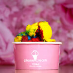 Close-up of a multicolor ice cream scoop (yellow, red, blue, green) in a pink paper cup with a pink, blurred background.