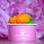 Close-up of a pink cup holding bright orange and green ice cream scoops with a purple blurred background, droplets on the cup.