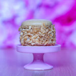Macaron covered with toasted coconut flakes on a pink cake stand, with a blurred purple background.
