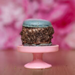 Blue macaron perched atop a crumb-covered chocolate dessert on a pink cake stand with a pink blurred background.
