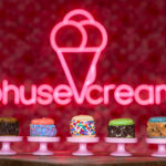 Row of colorful macarons on pink stands with a pink neon ice cream cone sign in the red-speckled background to the back.