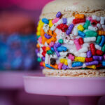 Close-up of a beige macaron with white filling, coated in colorful rainbow sprinkles on a pink pedestal.