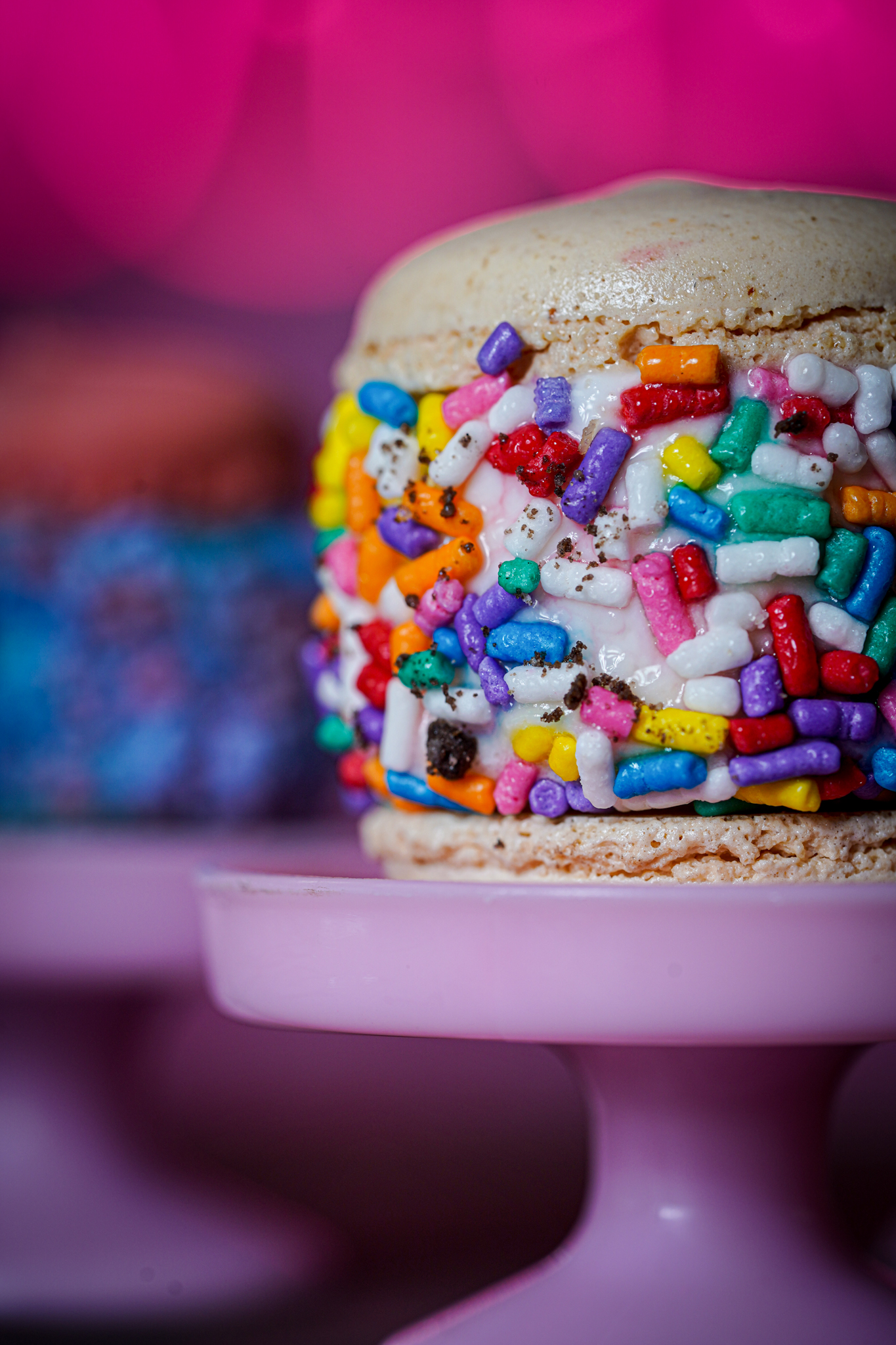 Close-up of a beige macaron with white filling, coated in colorful rainbow sprinkles on a pink pedestal.