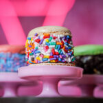 A donut coated in white icing with colorful rainbow sprinkles on a pink cake stand against a pink background.