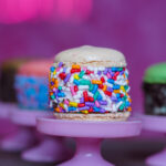 Vanilla donut with rainbow sprinkles on a pink pedestal against a purple background.